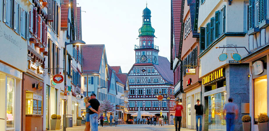 Eine Fußgängerzone in einer beleuchteten, dämmrigen Stadt. Geradeaus ist die Kirche und rechts sowie links sind Ladengeschäfte. In der Fußgängerzone sind ein paar vereinzelte Menschen unterwegs. 