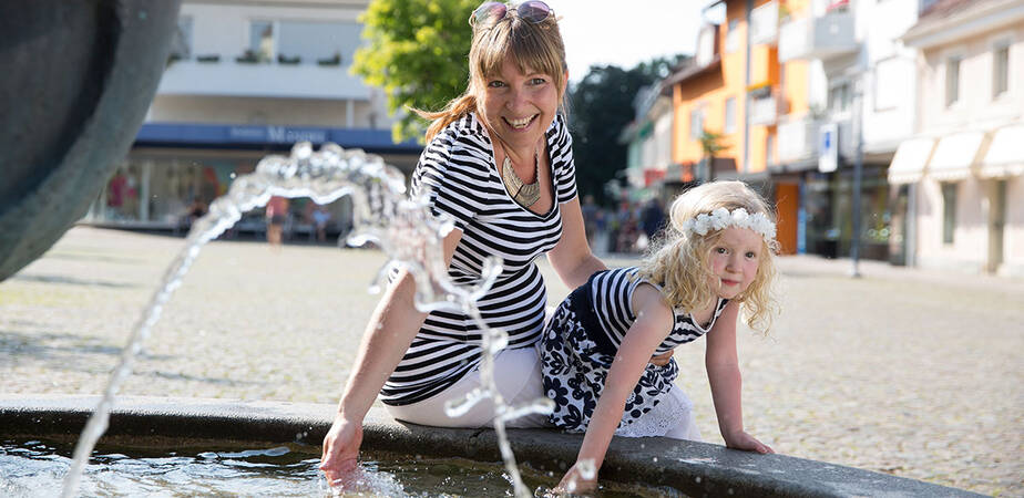 Eine Mutter mit ihrer kleinen Tochter, sie sitzen auf dem Rand eines Brunnens und halten ihre Hände in das Wasser. Im Hintergrund sind Ladengeschäfte der Stadt. 